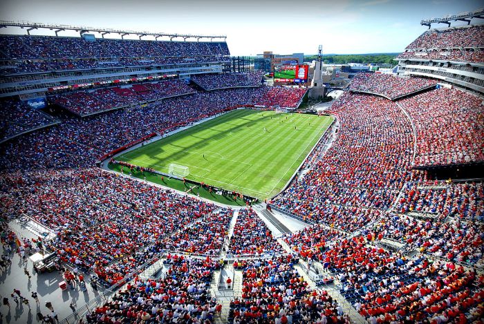 A large crowd fills Gillette Stadium around a soccer field. Players are on the field and a screen is above the stands. Most of the field and two-thirds of the spectators are in the bright sunshine.