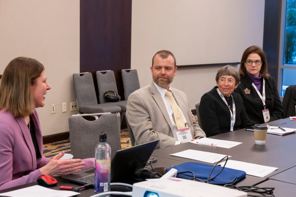 Victoria Sheehan speaks while Bastian Schroeder, Katie Turnbull and Ann Brach sit facing her at a table with a laptop, papers and a cup placed in front of them.