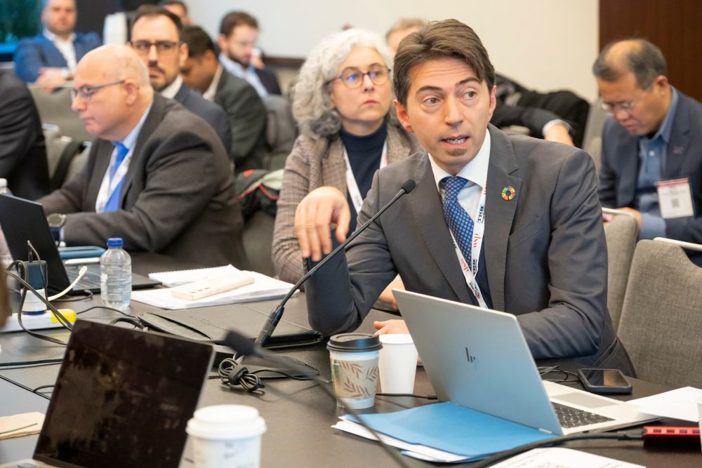 Biaggio Ciuffo, in formal attire with I D card, sits at a table and speaks into a microphone at the International Subcommittee meeting, with a laptop, papers, and a drink on the table and people sit behind and beside him.