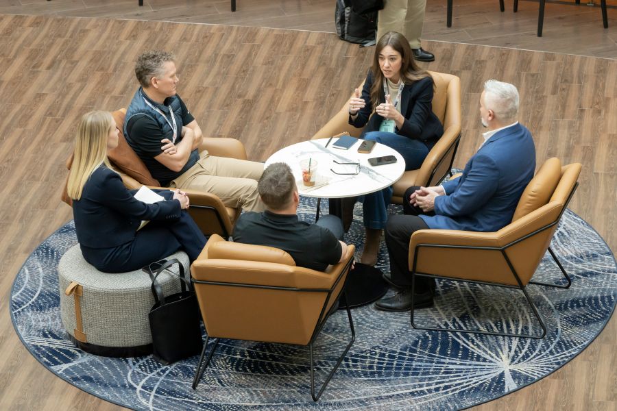 Five T R B Annual Meeting attendees sit on chairs around a table in an indoor space. Katherine Hunt speaks and the others listen.