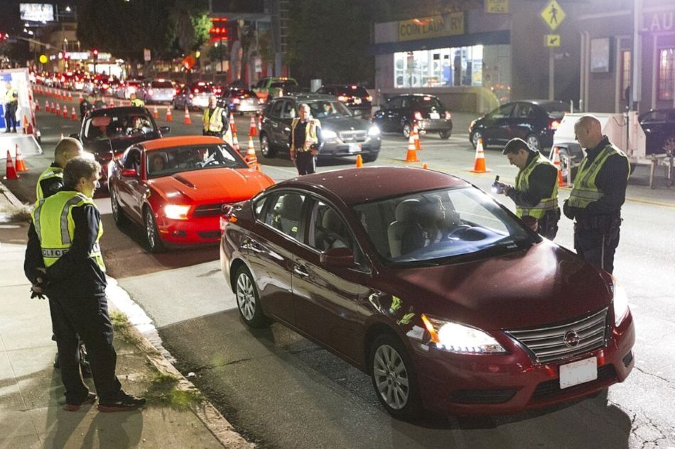 Police officers in Los Angeles check drivers at a sobriety checkpoint on a city street at night. Cars stop in lanes marked by traffic cones as officers speak with drivers and review documents.
