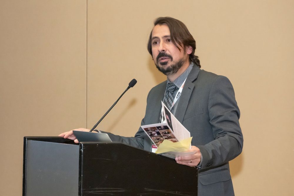 Samer Hamdar stands at a podium and speaks into a microphone. He holds a Hani S Mahmassani memorial service booklet.