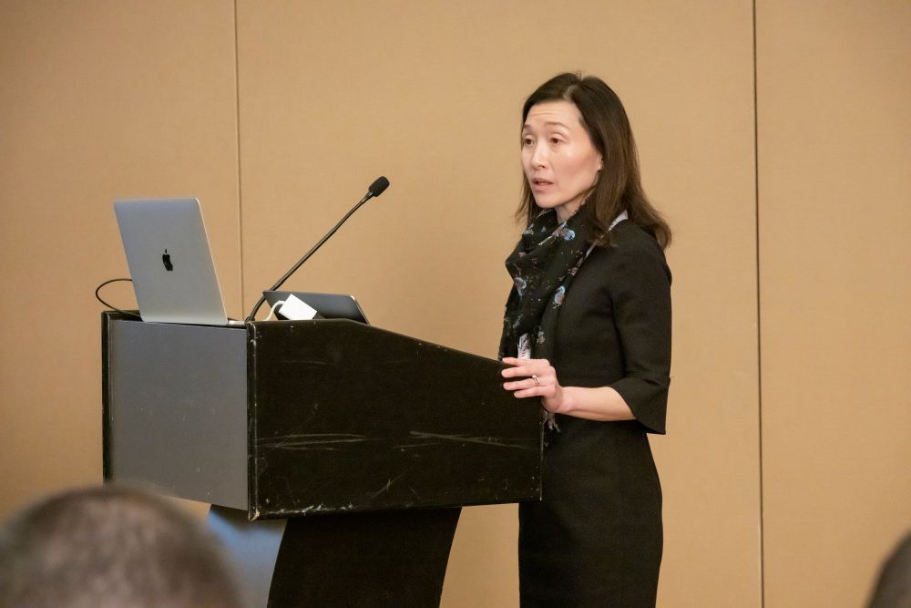 Sue Ahn stands at a podium and speaks at Hani S Mahmassani's memorial service. A laptop and a microphone are on the podium.