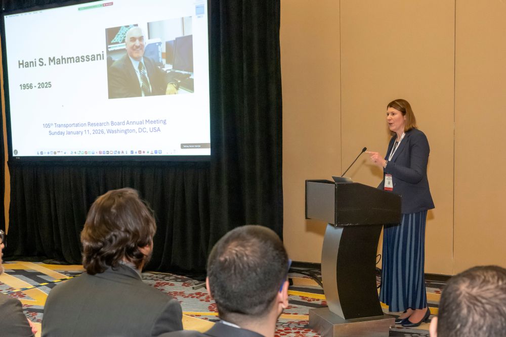 Victoria Sheehan, in formal attire, stands at a podium and speaks. A screen shows Hani S Mahmassani, 1956 to 2025. People sit and listen.