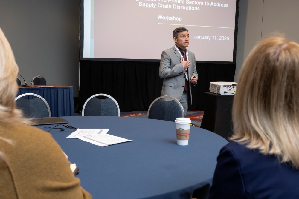 Mario Monsreal, in formal wear, stands and speaks in front of a screen during a workshop on addresssing supply chain disruptions while people sit at a table with papers and a cup.