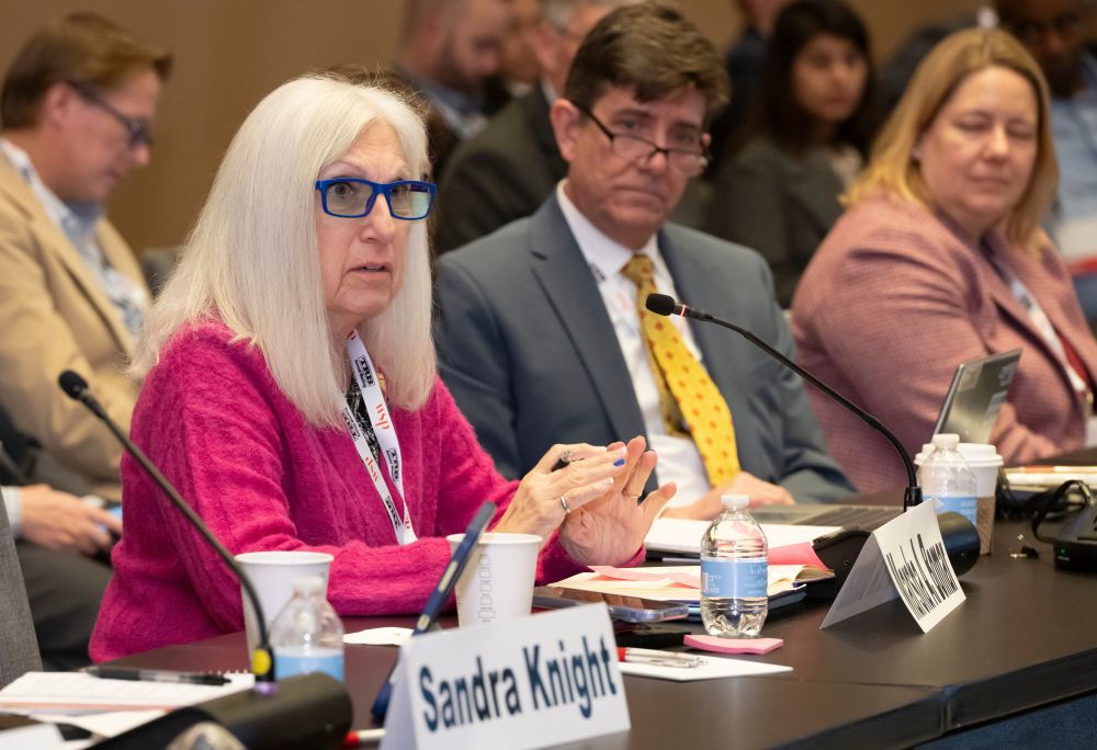 Marsha Anderson Bomar speaks while seated at a table with a microphone, papers, and a water bottle, as others sit beside her listening.