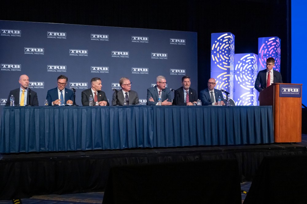 Derek Barrs speaks at a panel while Owen Morgan stands at the lectern. Other U S Department of Transportation administrators sit at the panel watching on.