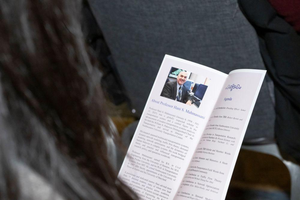 A person holds and reads a memorial service booklet. The booklet shows a photo and text about Professor Hani S Mahmassani and an agenda.