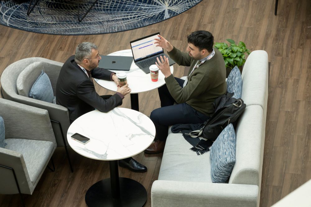 Two T R B Annual Meeting attendees sit at a table in an indoor space and talk while looking at a laptop, with beverages on the table and a backpack placed on a seat nearby.
