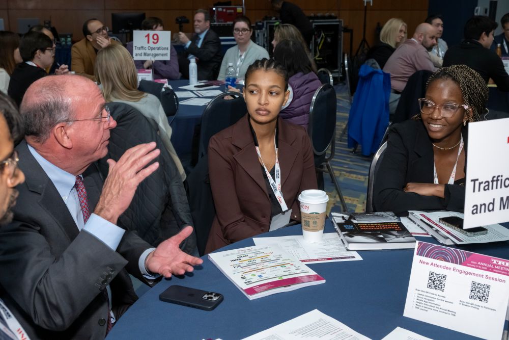 Larry Head speaks with Tsigereda Rose Mossie and Angel Angie Lucian at a round table. Papers, a phone, and a coffee cup rest on the table.
