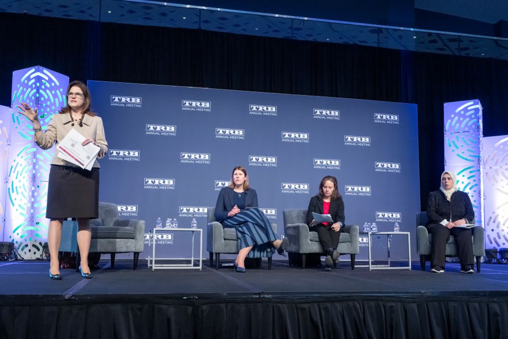 Ann Brach stands on a stage and speaks while holding papers, with Victoria Sheehan, Tara Cavalline, and Niloo Parvinashtiani seated on chairs behind her.