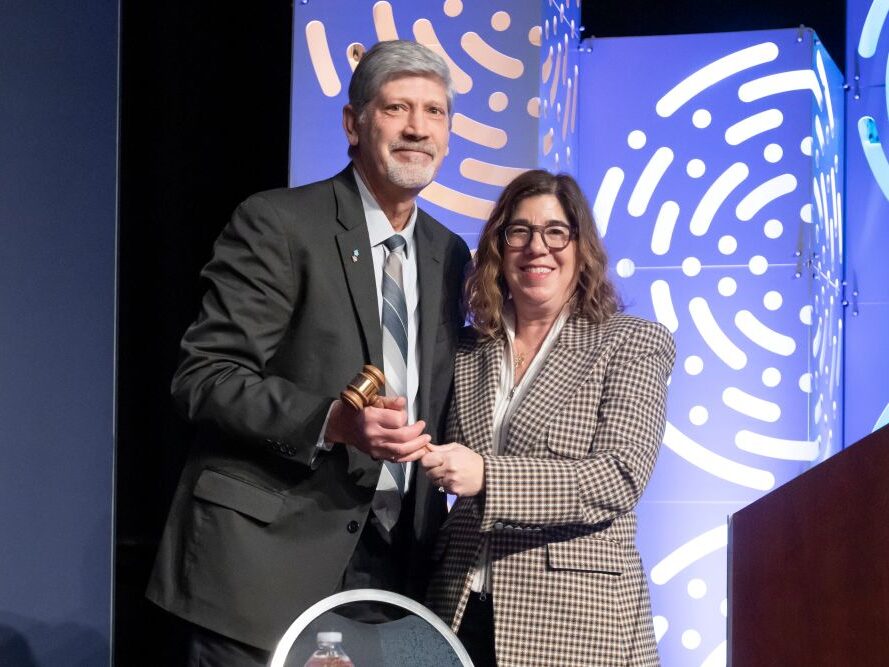 Passing the Gavel Incoming TRB Executive Committee chair Joel Jundt and 2025 chair Leslie Richards, both in formal attire, stand on a stage and hold a gavel together, smiling.