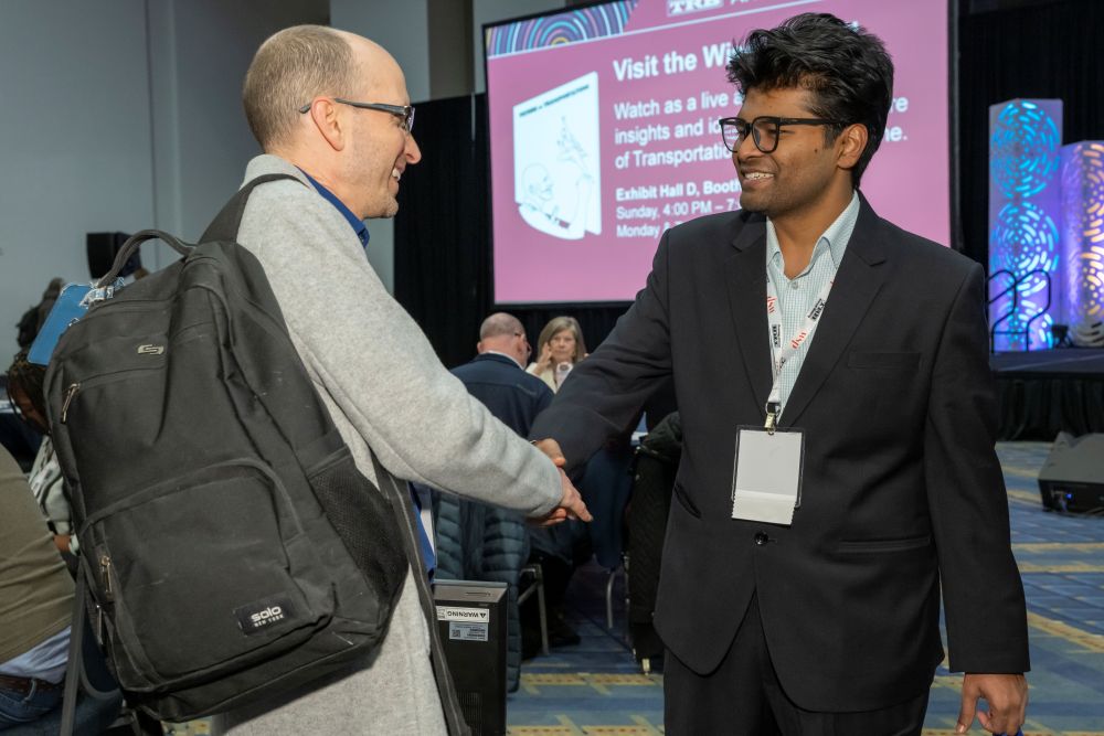 Peter Hylton and Muhammad Shajid shake hands in an indoor hall with a screen behind. Muhammad Shajid wears formal attire and Peter Hylton man wears casual clothing with a backpack.