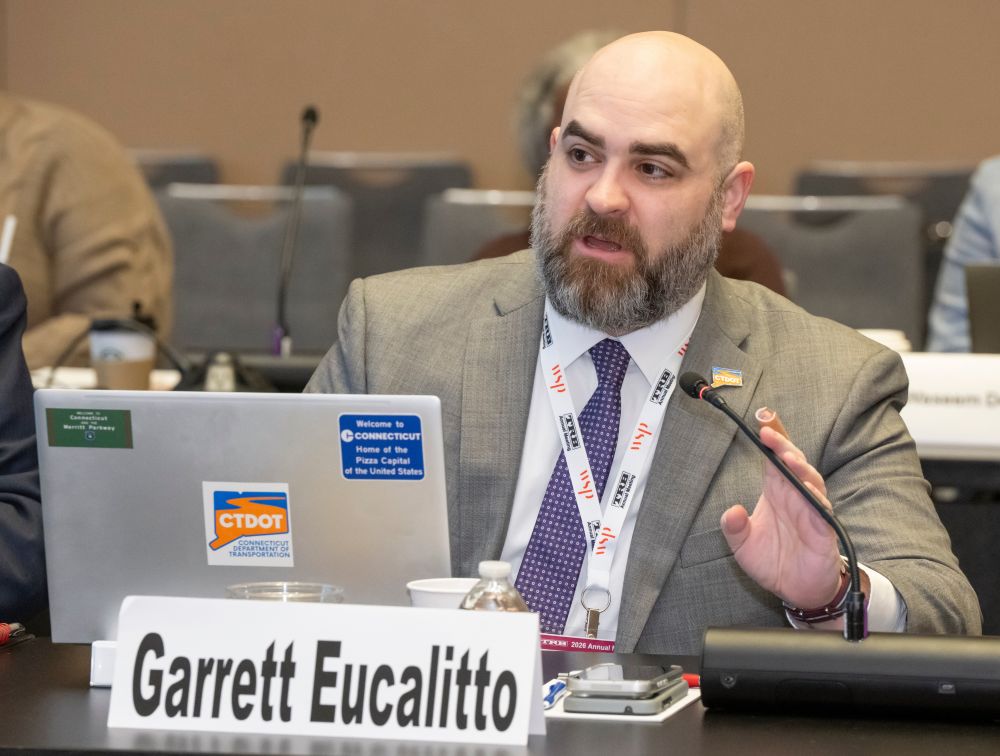 Garrett Eucalitto, in formal attire, sits at a table with a laptop and a name placard. A water bottle, a cup, and a microphone are on the table.