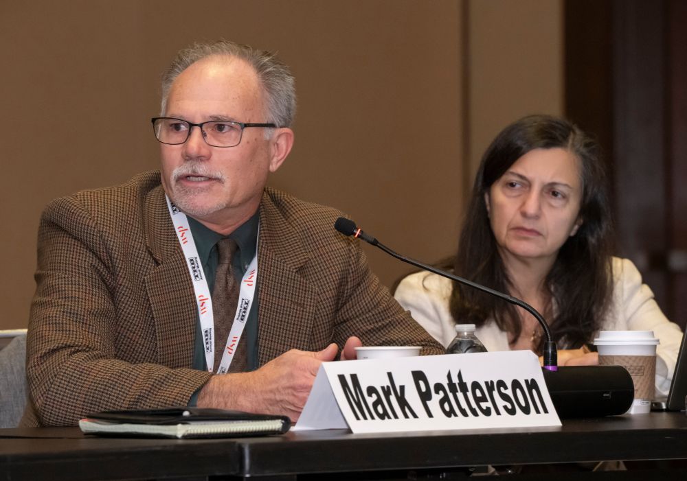 Mark Patterson speaks into a microphone while seated at a table with a notebook and name placard, as another participant sits beside him listening.