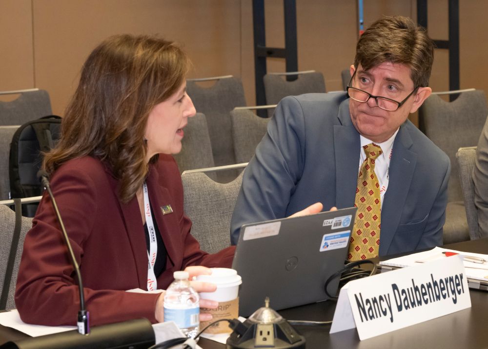 Nancy Daubenberger speaks while Joey Hartmann looks toward her, seated at a table with a laptop, papers, a cup, and a name placard in front.