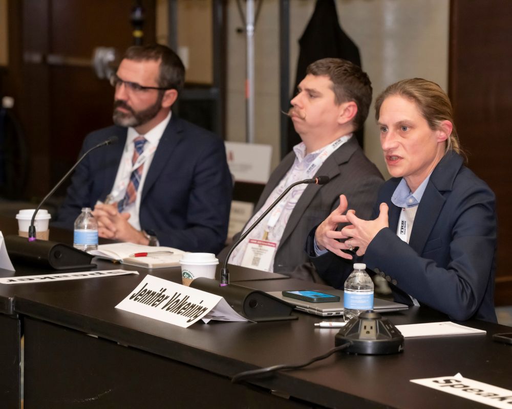 Jennifer Volkening speaks at the T R B Executive Committee policy panel while Gregory Ciparelli and Benjamin McCulloch sit beside her listening, with microphones, water bottles, and devices placed on the table.