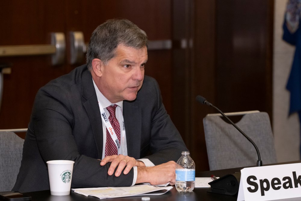 Tom Menzies sits at a table and speaks into a microphone, with a water bottle, cup, and papers placed in front of him.