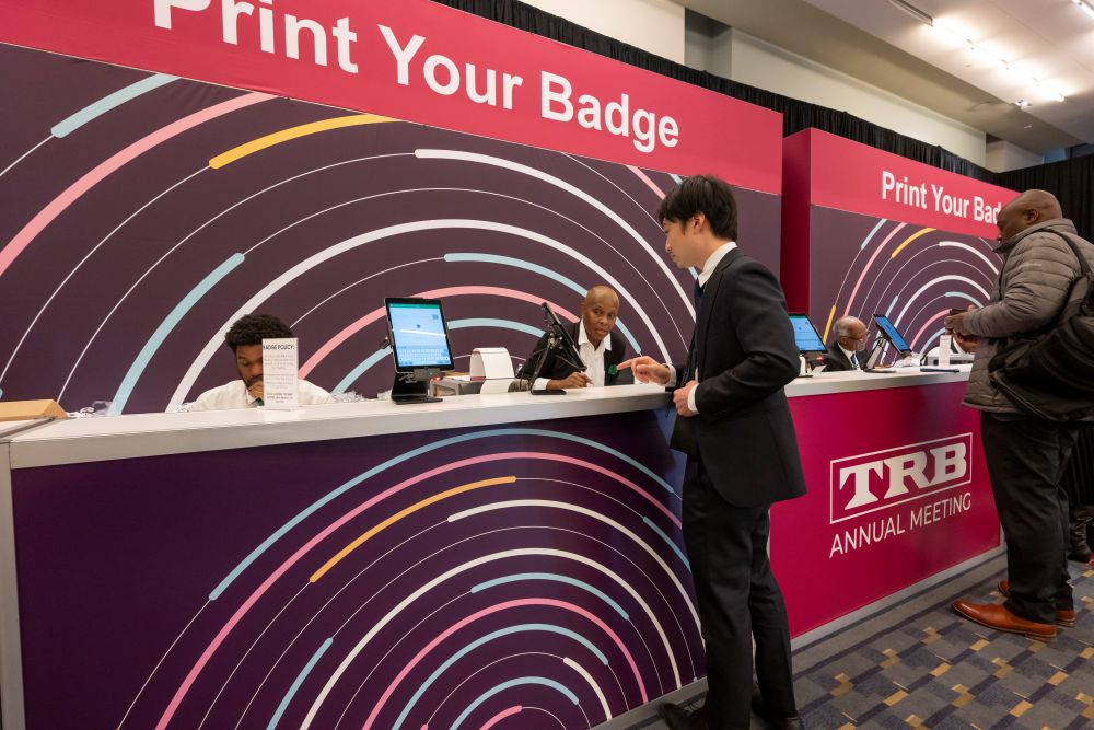 People stand at the T R B Annual Meeting registration counters to print badges. Staff work at computers behind the counters.