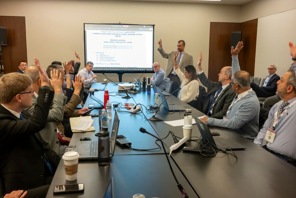 A group of commttee members sit around a table in an indoor room and raise their hands while Bastian Schroeder stands and leads the session near a screen with text.