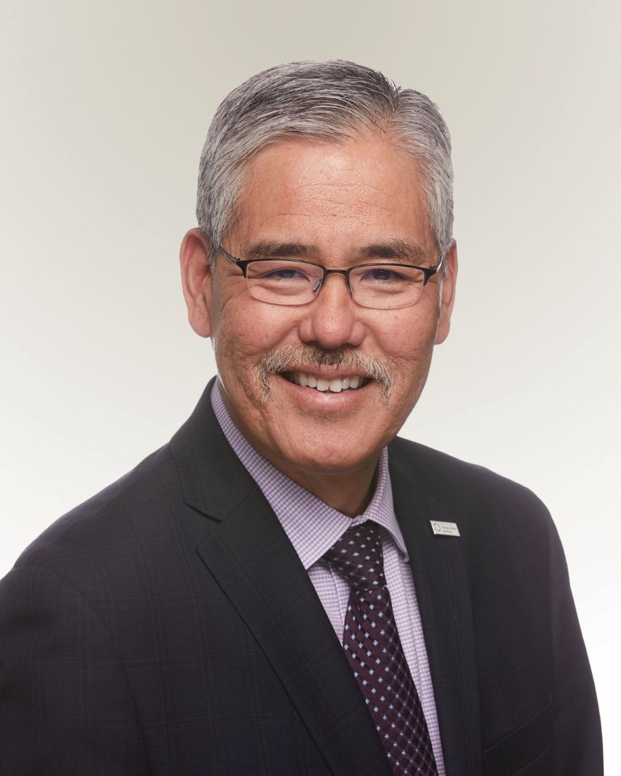 Randell Iwasaki, in formal wear with glasses, faces the camera and smiles.