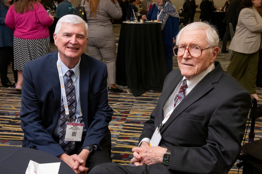 Wayne Kittelson and Thomas Deen sit together smiling, with attendees and tables arranged in the background.