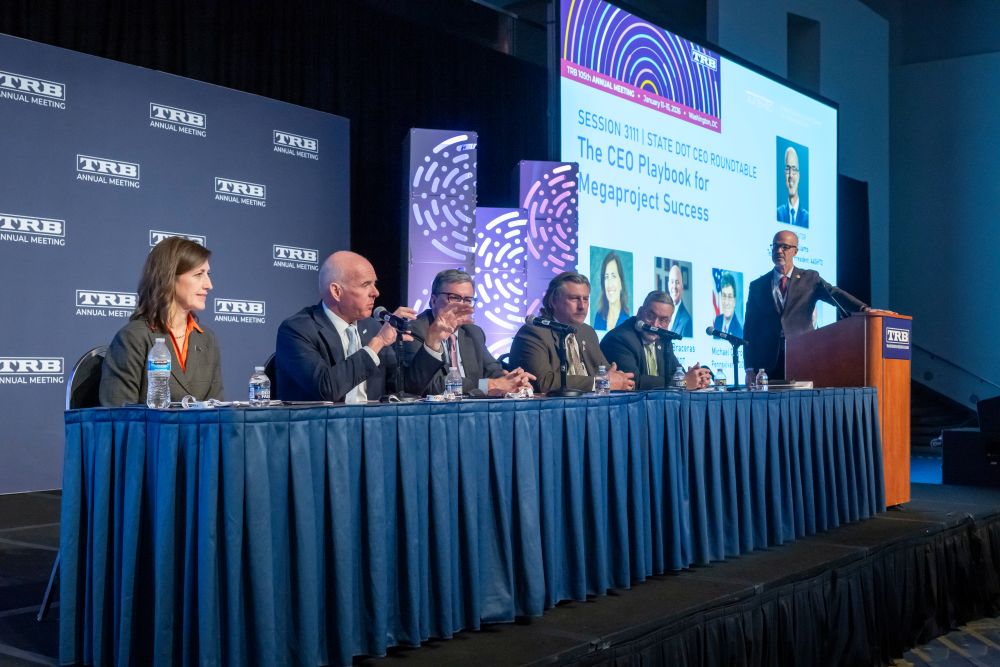 A panel of state D O T chiefs, in formal wear, sit at a table on a stage while Carlos Braceras speaks into a microphone and Marc Williams stands at a lectern, with a screen behind them.