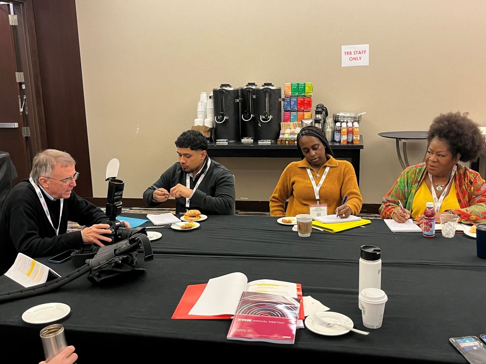 Photographer Cable Risdon with camera sharing notes with three staff photo assistants at table in the T R B staff room. There is a coffee and tea station behind them and cups, juice drinks, paper plates with food, and a T R B Annual Meeting program on the table.