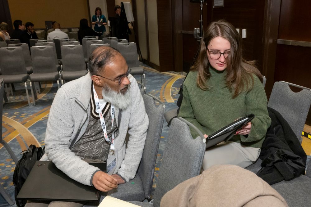Rehan Hyder looks toward Emma Habosky as she holds a document while seated among arranged chairs.