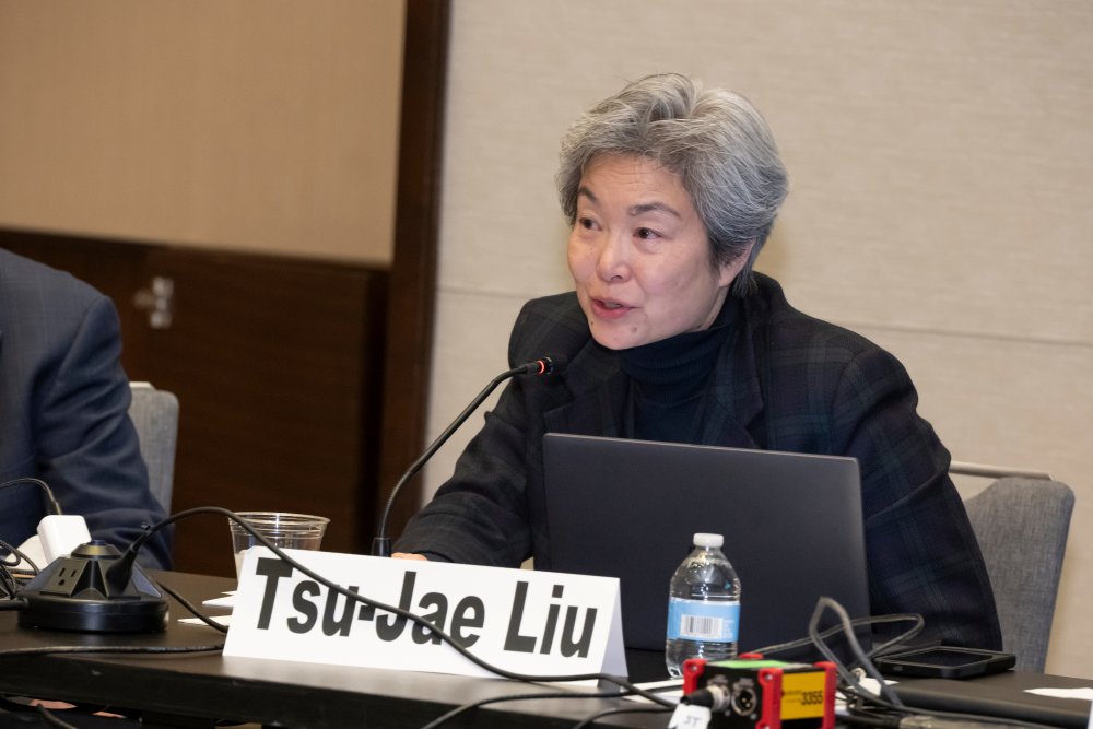 Tsu-Jae Liu speaks into a microphone while sitting at a table with a laptop, a water bottle, and equipment placed in front of her.