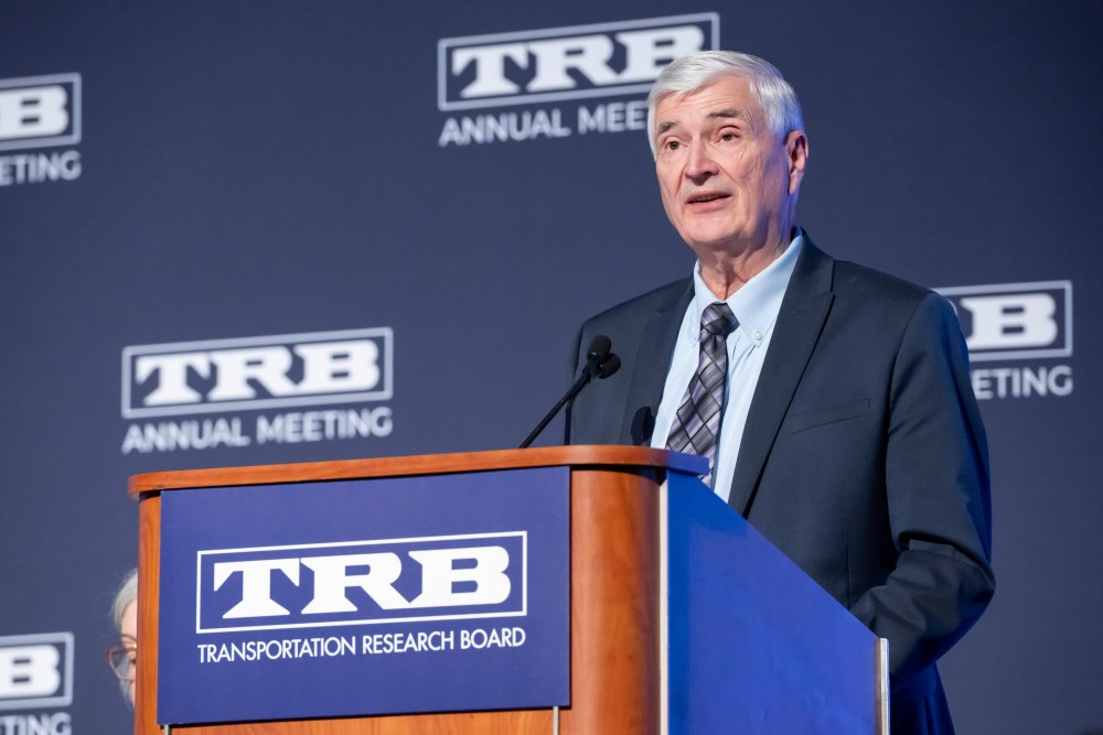 Wayne Kittelson in formal wear stands at a podium and speaks, with a backdrop showing T R B Annual Meeting text and text on the podium reading T R B Transportation Research Board.