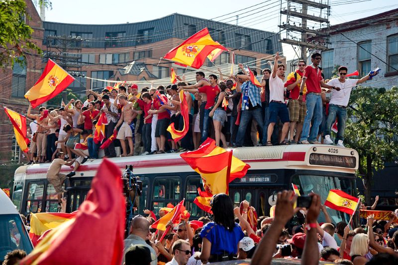 A group of World Cup soccer fans stand on top of a bus and wave Spanish flags. More people stand around the bus and hold flags in a crowded street.
