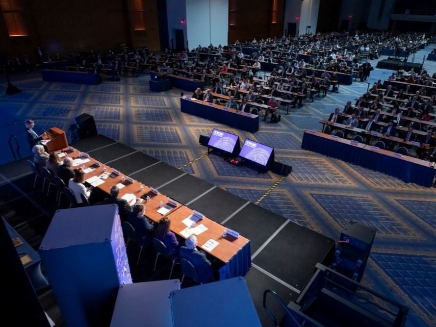 Keynote Speaker and Audience.2 A large ballroom with many people seated at tables facing a stage where one speaker stands at the lectern and panelists sit at a long table.