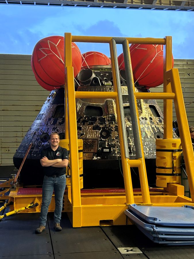 A NASA engineer stands with his arms crossed, smiling, in front of the cone-shaped crew capsule . The capsule has 3 large inflated balloons at the top of its nosecone and is resting on a platform with a tall support structure.