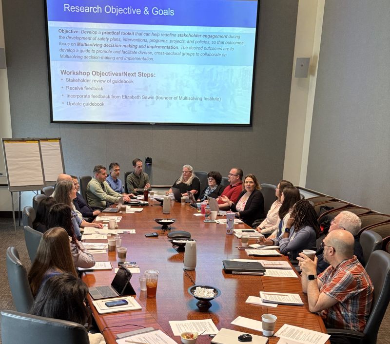 A group of workshop participants in T R B's Behavioral Traffic Safety Cooperative Resarch Program Project B T S 24 sit around an oval conference table with papers and laptops in front of them. A slide on the projector screen above the head of the table reads: Research Objectives and Goals and lists workshop objectives and next steps.