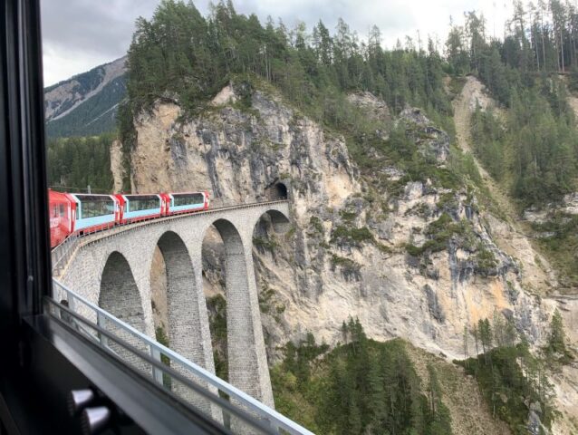 A Swiss train crosses a tall arched stone viaduct and enters a tunnel in a rocky forested hillside.
