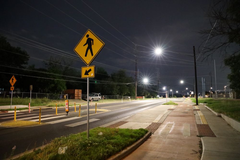 A nighttime crosswalk has pedestrian signs, bollards, bright streetlights, sidewalks, and raised pavement pads along the road.