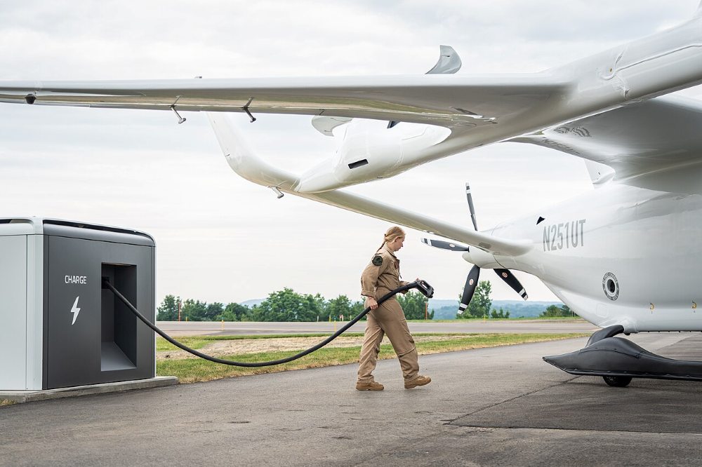 A person in a flight suit carries a charging cable from a cubic charging unit toward a circular charging port beneath the wing of an electric aircraft.