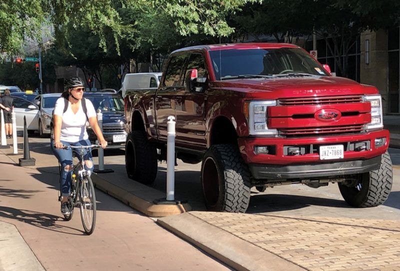 A bicyclist rides on a bollard-protected bike lane beside a large, raised pickup truck with oversized tires on a city street with cars, and trees nearby.
