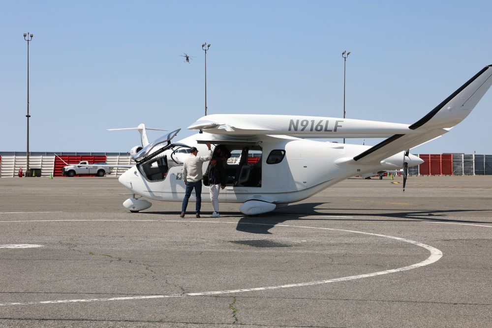A small electric plane labeled N 916 L F sits on the tarmac at JFK International Airport with its door open while the pilot and a passenger stand beside the door.