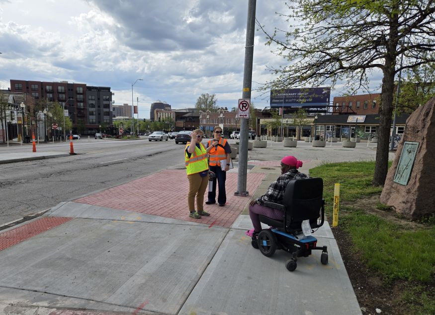 Two workers wearing safety vests talk with a wheelchair user on a paved sidewalk beside a street. Tactile paving, a signpost, and buildings are nearby.