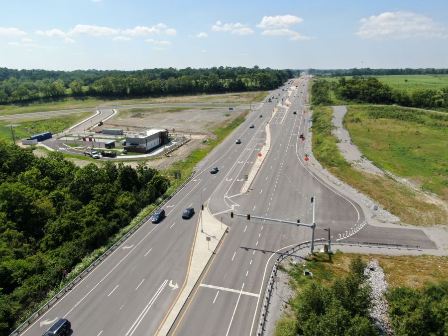 An aerial view shows a wide road intersection with J-turn or U-turn lanes, vehicles, signals, and open land nearby.