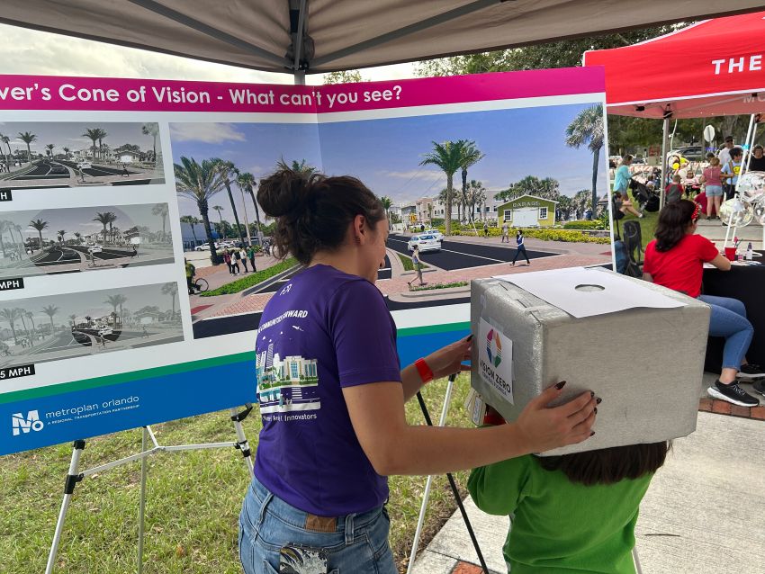 At an outdoor event, a person helps a child use a vision activity box by placing it on the child’s head. A display board about driver visibility is in front.