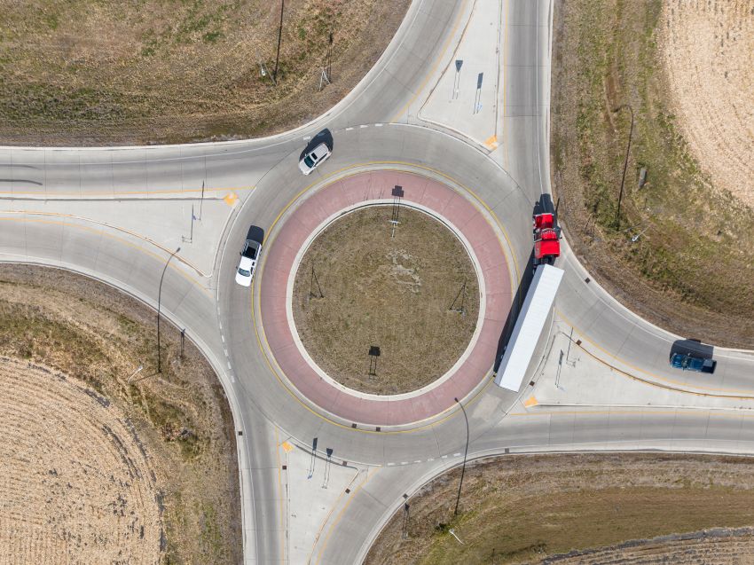 An aerial view shows a roundabout with separated traffic lanes, a freight truck and several vehicles, marked crossings, and open fields around the roads.