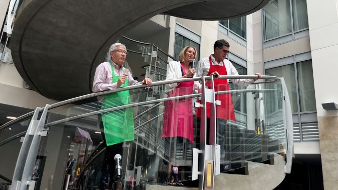 Three people wearing apron smocks stand on a spiral staircase. The man on the left holds a fudge bar. The woman in the center is speaking into a hand-held microphone. The man on the right has both hands on the railing and is looking downwards.