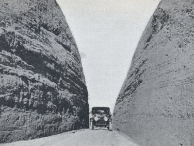 An early automobile travels through a narrow roadway cut between two tall rock walls.