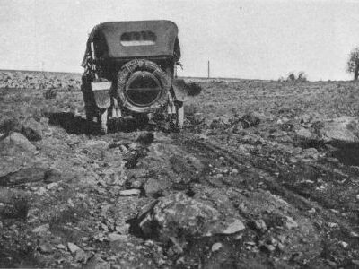 An early automobile travels over a rough rocky road with deep ruts across open land.