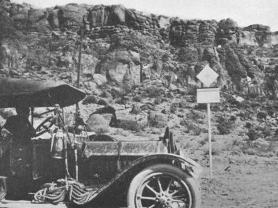 An early automobile with a rope and lantern is parked beside a rocky hillside with a roadside sign nearby.