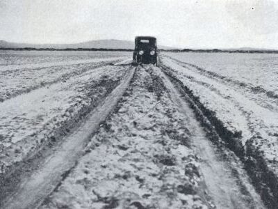 An early automobile travels along a deeply rutted road across flat open land. Uneven tracks extend from the foreground toward the automobile.