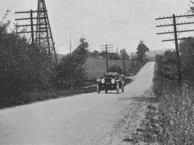 A rural road extends through trees and fields with utility poles on both sides and an early automobile traveling near the center.